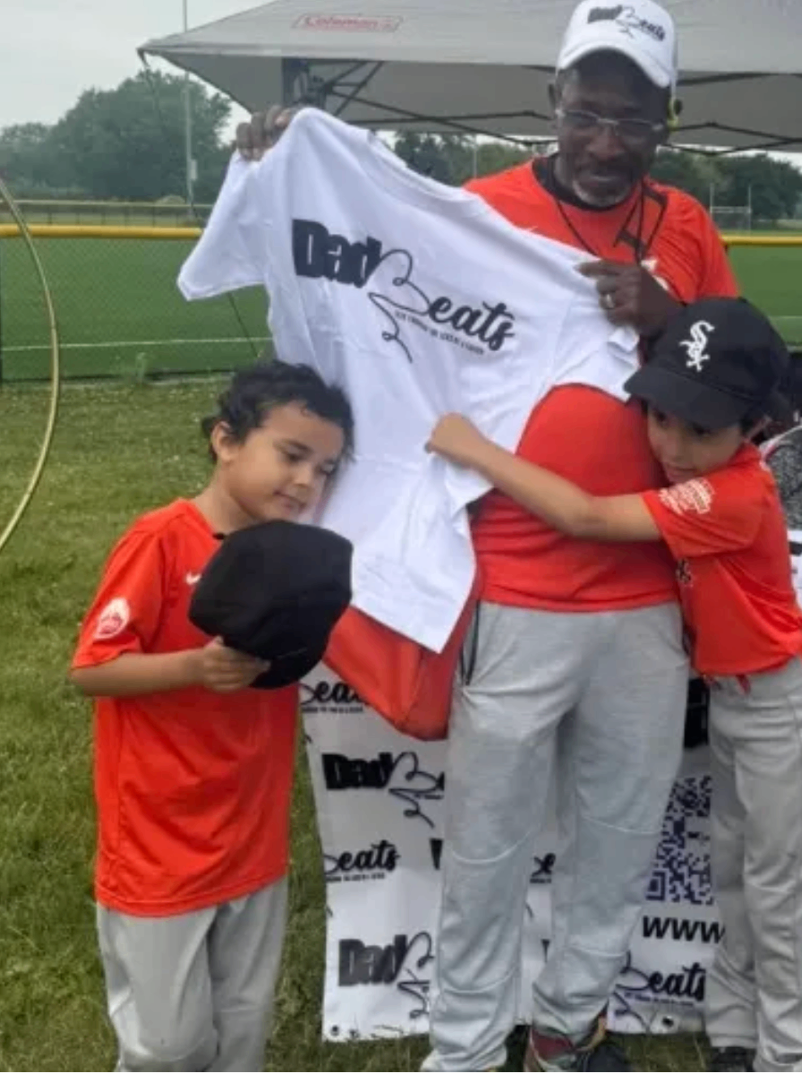Grandfather holding up a Dad-Beats shirt while two young boys hug him at the competition booth